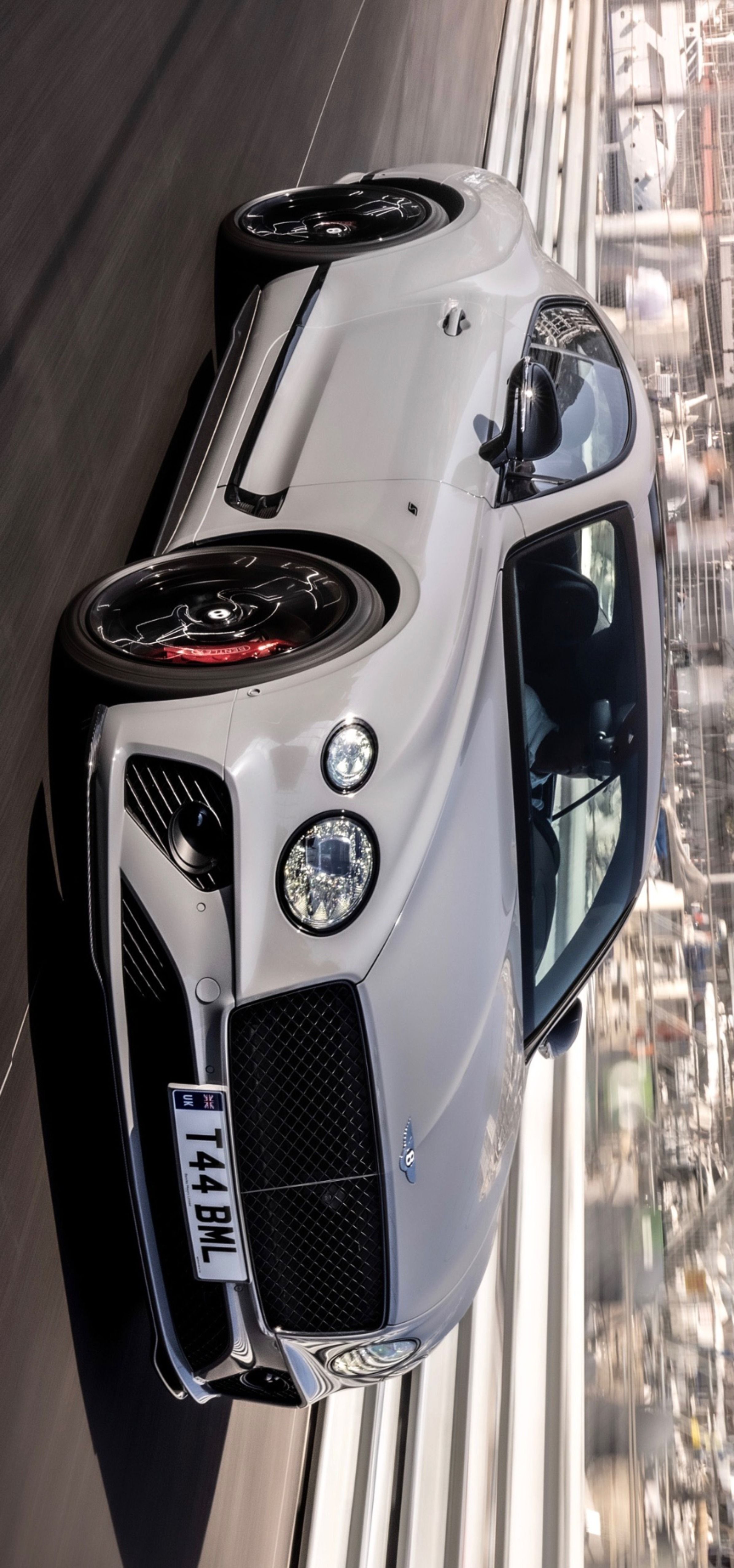 the front end of a white sports car driving down a road with buildings in the background